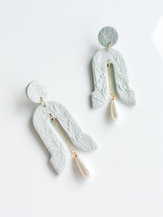 The Gloria earrings by Agave Designs feature light gray textured polymer clay with floral patterns, gold chains, and white teardrop pearls on sterling silver hooks, shown on a white background.