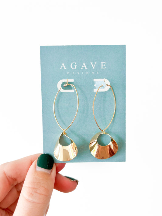 A hand with dark green nail polish holds a card labeled "Agave Designs," displaying the Halo Danglies—the stunning 53mm gold hoop earrings made from zinc alloy, featuring circular reflective pendants. The earrings are presented against a plain white background.