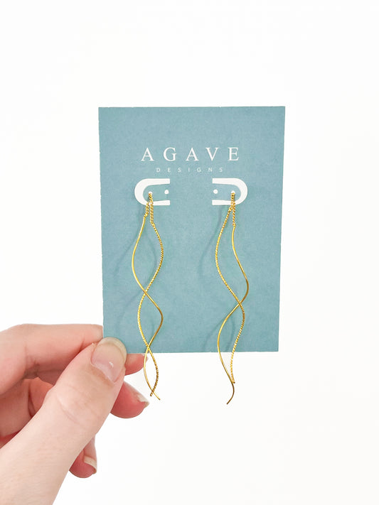 A hand displays a light blue card featuring the Gold Wave Tassels from Agave Designs, an elegant pair of 80mm twisted wire earrings with a wavy design in stainless steel.