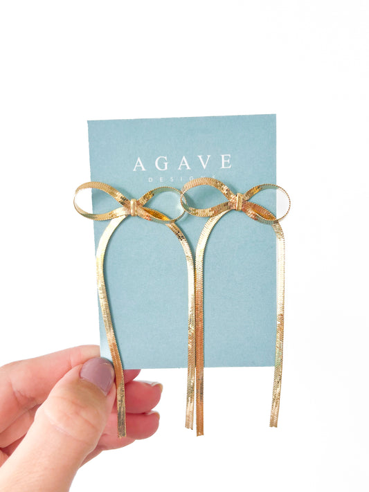 A hand holding a pair of Agave Designs "Big Bows" drop earrings attached to a blue Agave Designs card. The delicate gold earrings, made from Zinc Alloy, feature long, dangling ribbon strands and measure 90mm x 40mm. The background is plain white.