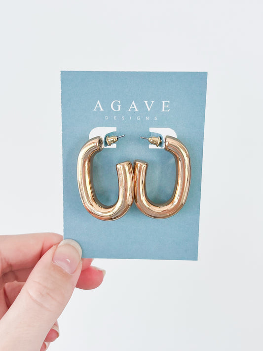 A hand displays a pair of elegant Chunky 1/2 Hoops crafted from zinc alloy, mounted on a blue card marked with the "Agave Designs" brand, set against a clean white backdrop.