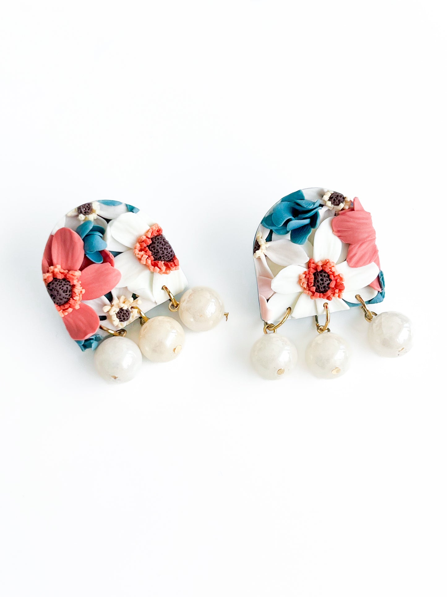 Introducing "Joy" by Agave Designs: A stunning pair of decorative polymer clay earrings with an arch shape, showcasing colorful floral designs in red, white, and blue. Each earring features three dangling round beads at the bottom, resembling off-white pearls. The background is a simple white hue.