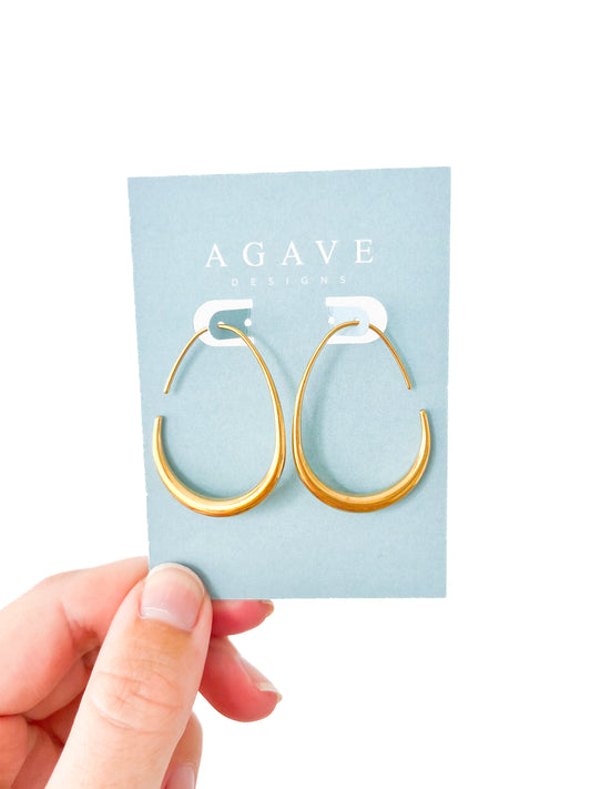 A hand holds a pair of 40mm x 28mm Oval Drops - Large gold hoop earrings displayed on a light blue card with "Agave Designs" written on it in white text. The earrings, made of polished stainless steel, feature a smooth, minimalist design with an open section at the top. The background is white.