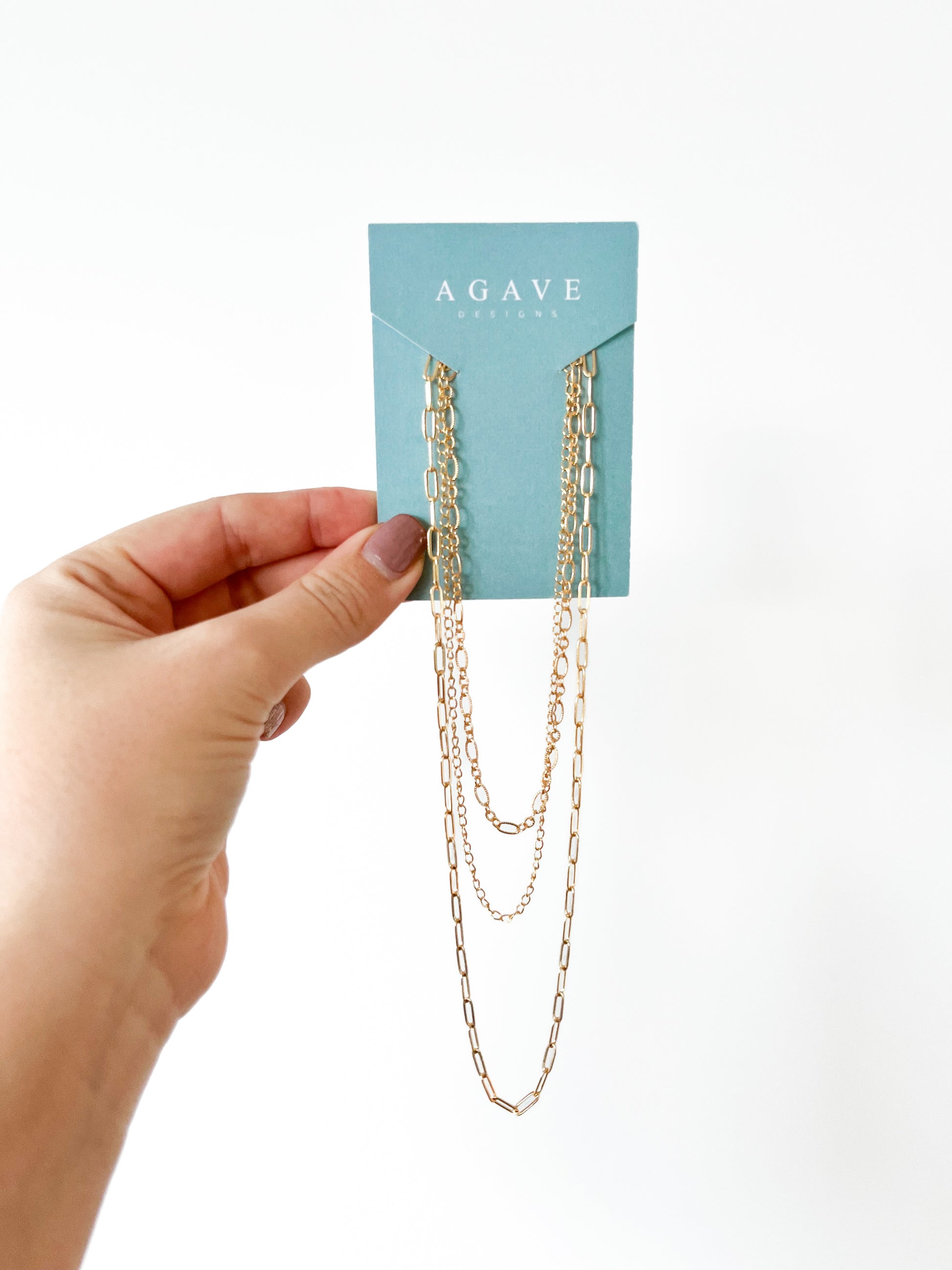 A hand with manicured nails is holding a turquoise card displaying layered gold necklaces. The card is branded with the name "Agave Designs." The 3 Tiered Necklace measures up to 55cm with diverse chain styles and lengths, hanging elegantly against a white background.