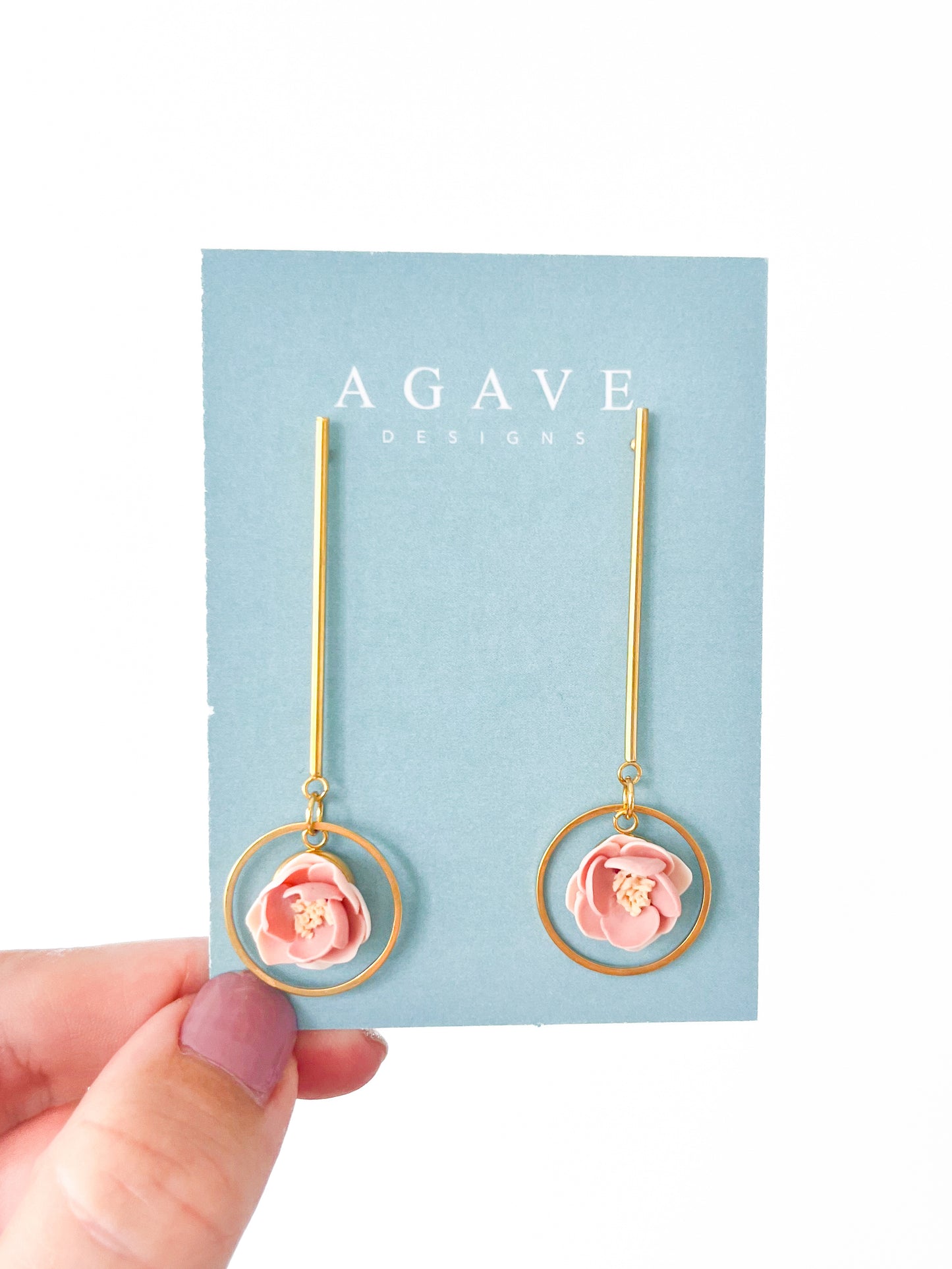 A pair of gold-tone Rose Drop Danglies featuring circular frames with small dusty pink and peach flowers inside. Each earring is crafted from gold plated stainless steel and is displayed on a light blue "Agave Designs" card held by a hand with purple nail polish.
