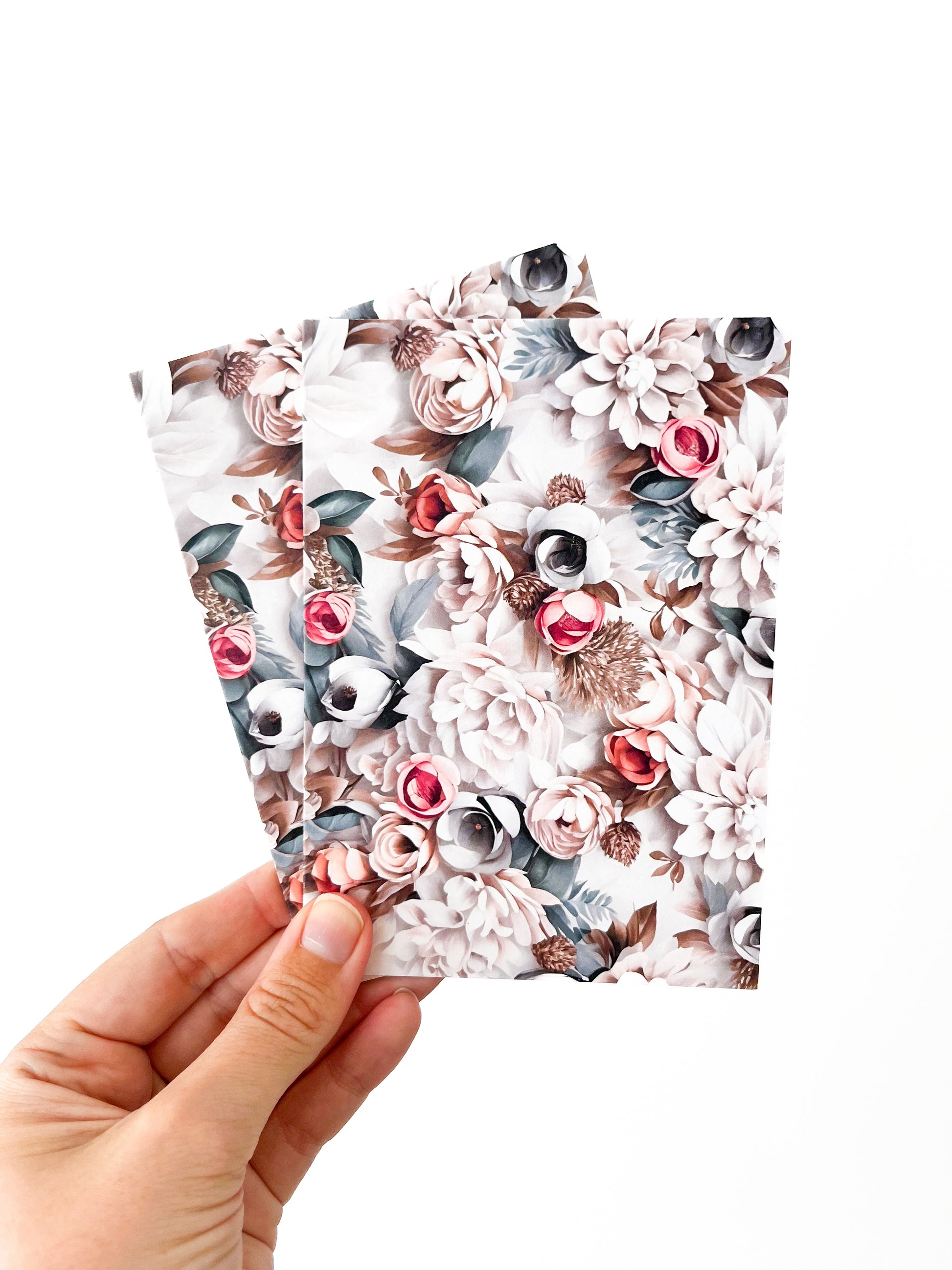 A hand holding three greeting cards from Agave Designs against a white background. The cards, housing exquisite polymer clay earrings, showcase a soft, delicate design featuring various flowers in shades of pink, white, and gray.