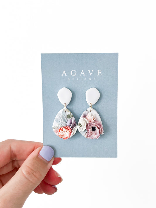 A hand with lavender nail polish holds a light blue card reading "Agave Designs," displaying Julianne sterling silver drop earrings with white tops and teardrop pendants featuring pastel floral designs.