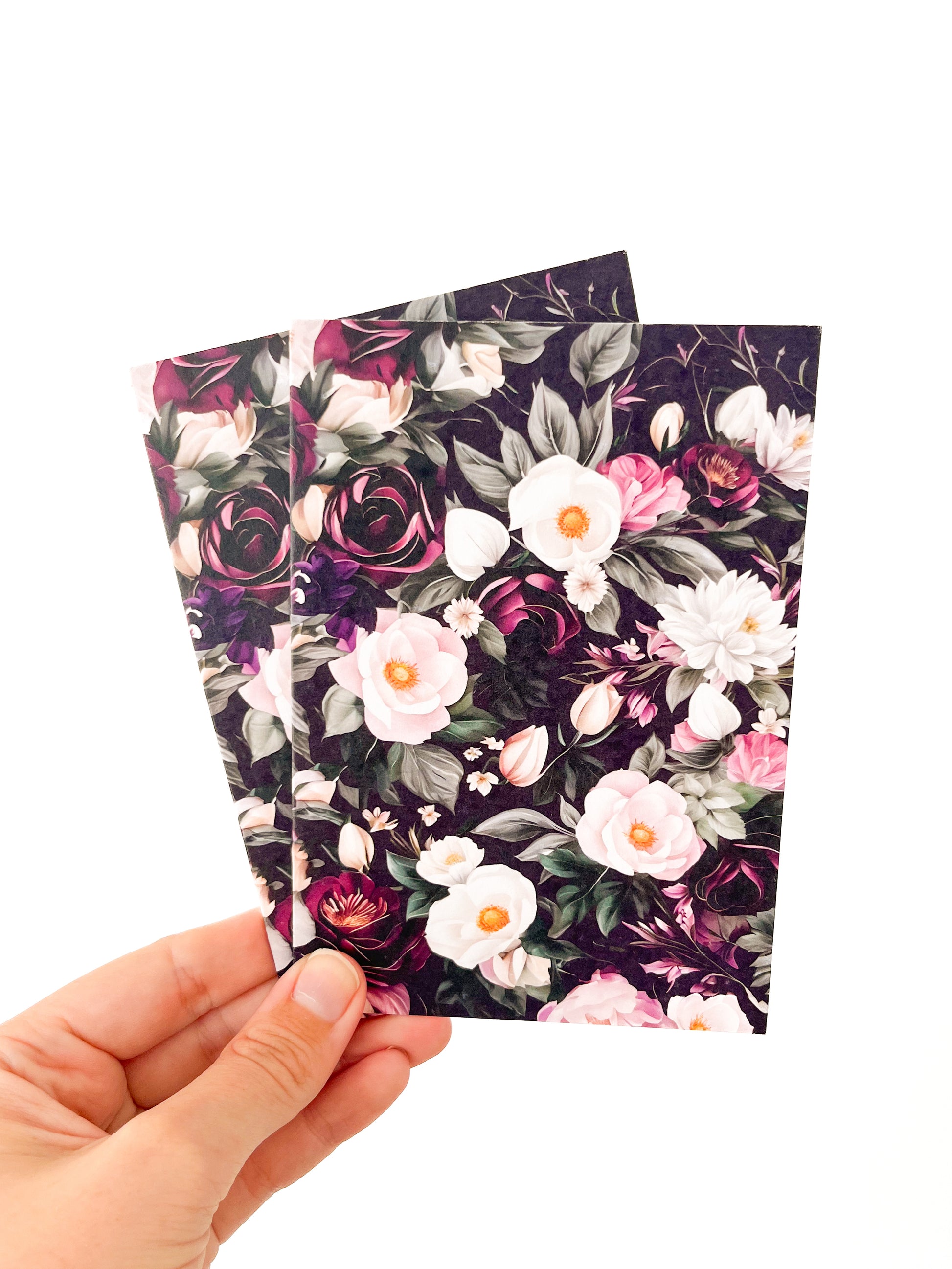 Two A5 greeting cards from Agave Designs, showcasing vibrant floral designs, are held in a person's hand against a white background. These greeting cards feature a variety of flowers, including white roses and pink blossoms, surrounded by dark green leaves and small white buds.