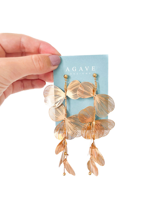 A hand with light purple nail polish holds a light blue card labeled "Agave Designs" showcasing a pair of Gold Petal Danglies, which are gold, leaf-shaped polymer clay earrings featuring multiple delicate leaf charms in a cascading design.