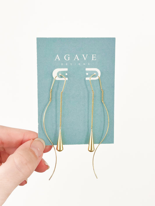 A hand is holding a teal card labeled "Agave Designs," showcasing the Long Water Drop Danglies, a pair of long, gold earrings made from zinc alloy with a sleek, modern design. These 90mm earrings feature a delicate chain and a cone-shaped pendant.