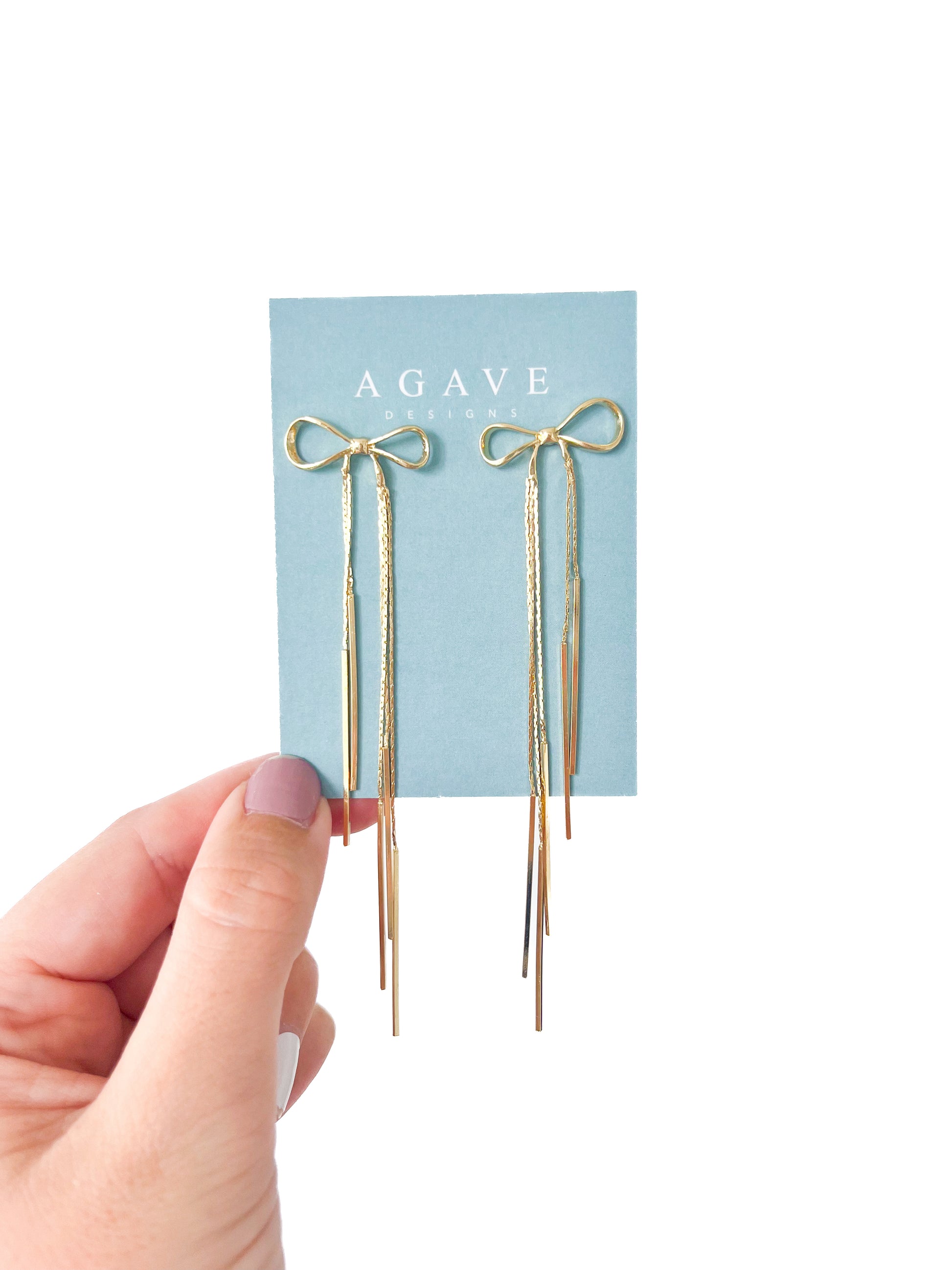 A hand with light pink nail polish holds a light blue card from "Agave Designs" showcasing the "Little Bow Danglies," exquisite gold bow-shaped earrings made from copper alloy. These earrings feature long, dangling strands and measure 110mm x 26mm, epitomizing minimalist sophistication with their thin and elegant design.