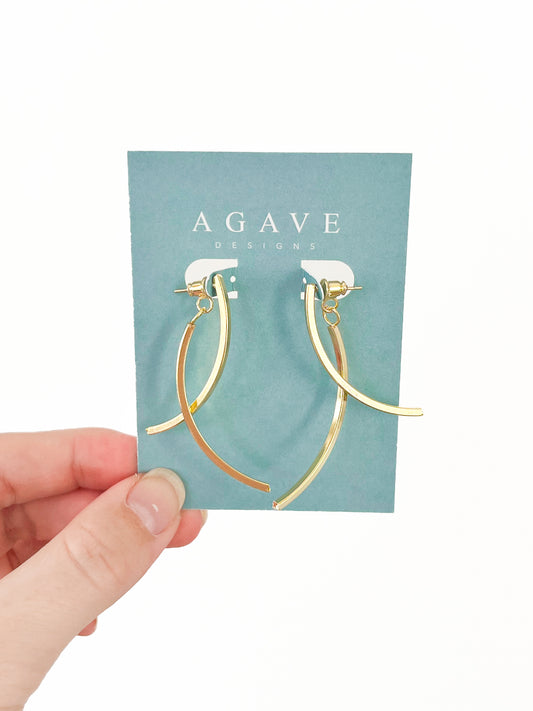 Hand holding a 53mm card from Agave Designs with the "Arc Drops," a pair of gold, curved earrings made from Zinc Alloy attached. These earrings showcase a sleek, modern design featuring two elegant hooks. The card is turquoise with white text.