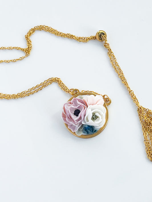 The Agave Designs Floral necklace is a gold plated adjustable piece with a round pendant featuring pink, white, and blue flower designs, shown on a white background.