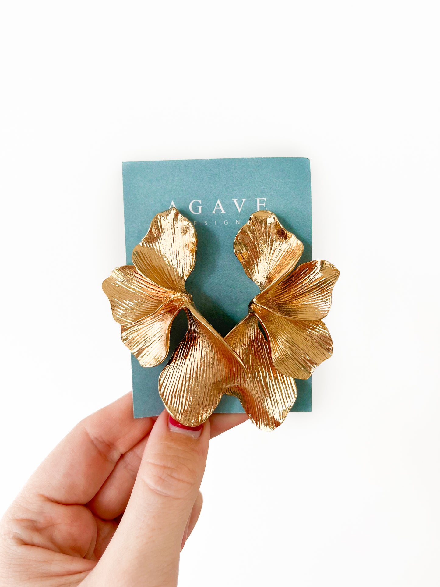 A hand displays a pair of Chunky Gold Petals earrings measuring 75mm x 50mm, shaped like leaves against a blue-green card labeled "Agave Designs." Crafted from zinc alloy, the intricate detailing of these stunning gold metallic earrings is accentuated against the plain white background.