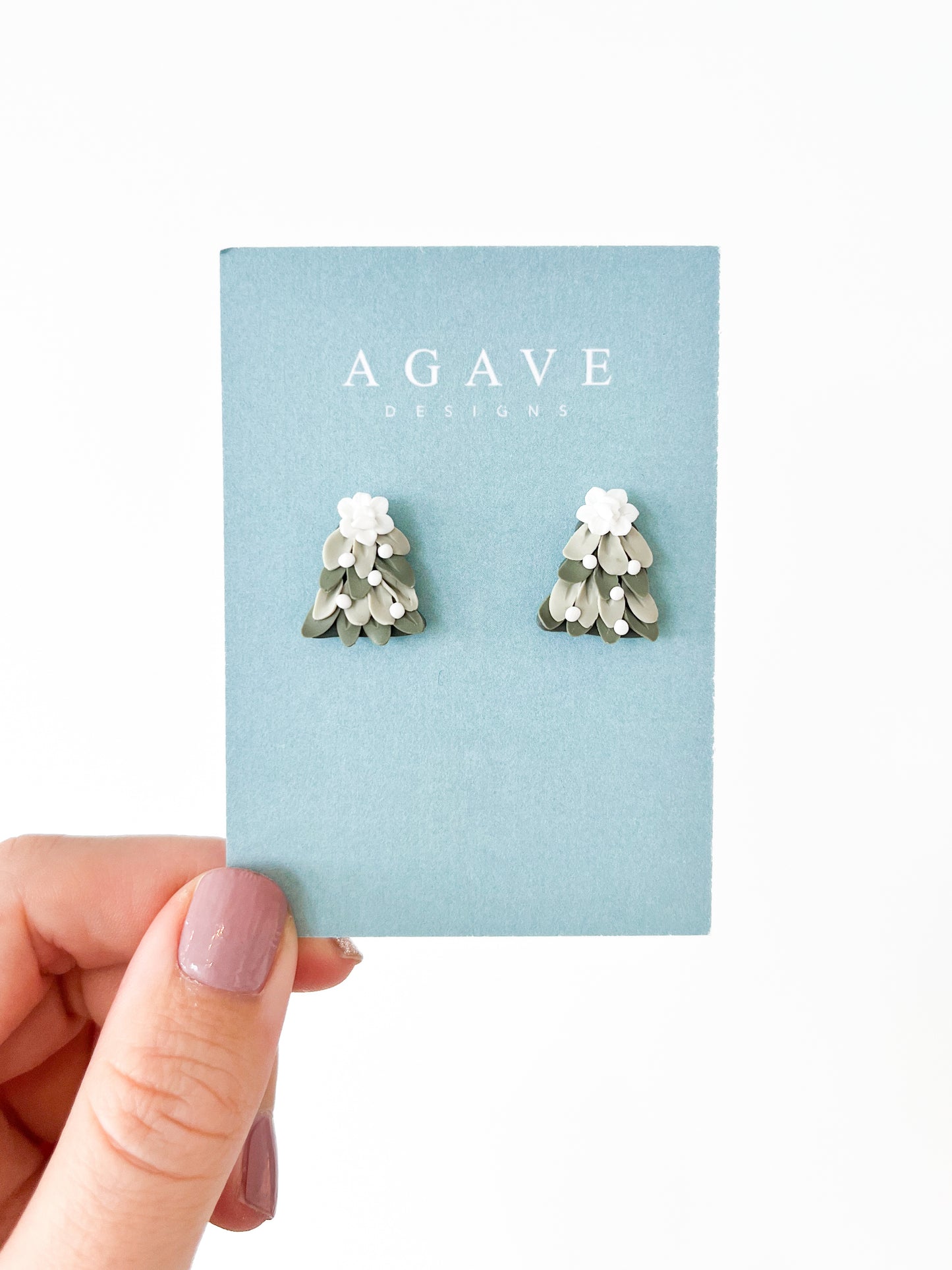 A hand displays a card labeled "Agave Designs" featuring handmade earrings called "Christmas Tree Studs." Crafted with sterling silver, these 20mm x 15mm beauties showcase layered greens and white flower designs, standing out against a light blue background.