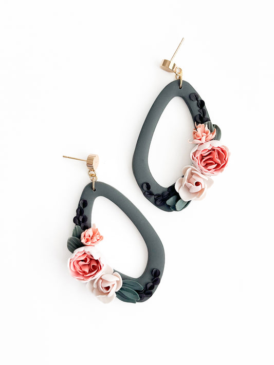 A pair of teardrop-shaped Harmony earrings by Agave Designs on a white background. The earrings are black with decorative floral accents in pink, white, and forest green hues, featuring roses and leaves. The gold-plated earrings have gold-tone posts and backings and measure 60 x 30mm.
