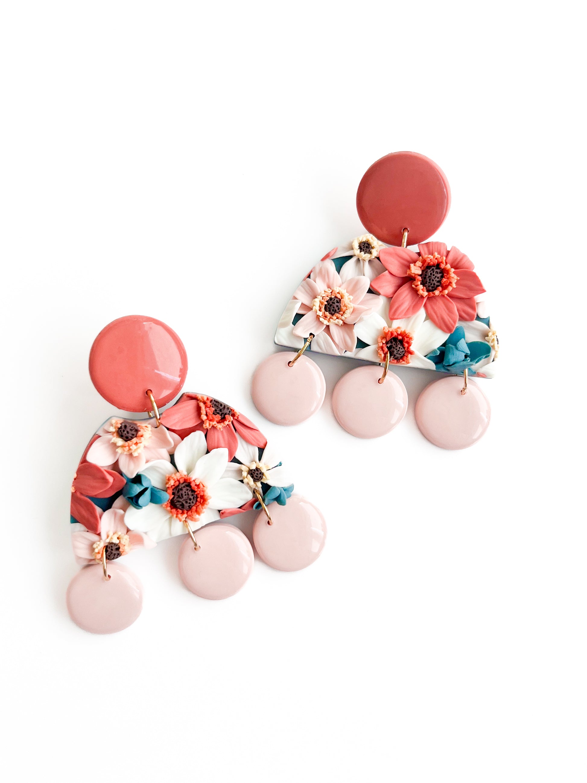 The Dottie earrings by Agave Designs are colorful polymer clay pieces featuring red, white, and teal flowers on a semi-circular base, adorned with large pink circles. This unique design combines vibrant florals and geometric shapes to create a bold and artistic accessory.