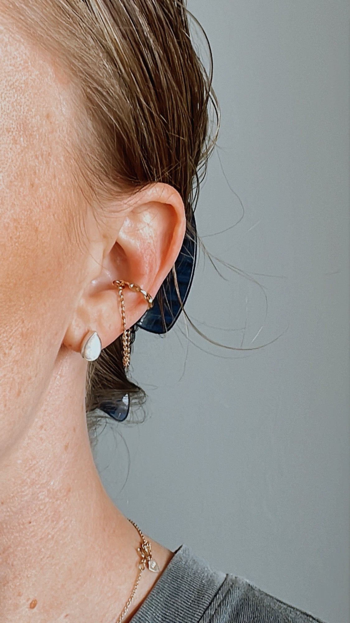 Close-up of a person's ear adorned with Agave Designs' Ear cuffs, featuring a pear-shaped stud and a decorative, twisted, coil-like design. With their hair pulled back and secured by a dark hair tie, the fair skin is highlighted beautifully. Polymer clay earrings add an artisanal touch against the plain gray background.