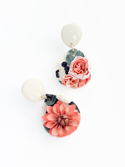 The Bonnie earrings by Agave Designs are a stunning pair of handmade accessories featuring white top studs and floral drop pendants. One earring boasts a detailed pink rose, while the other displays a vibrant coral dahlia, both beautifully accented with forest green leaves and small black details on a white background. These gold-plated earrings measure 40mm x 17mm.