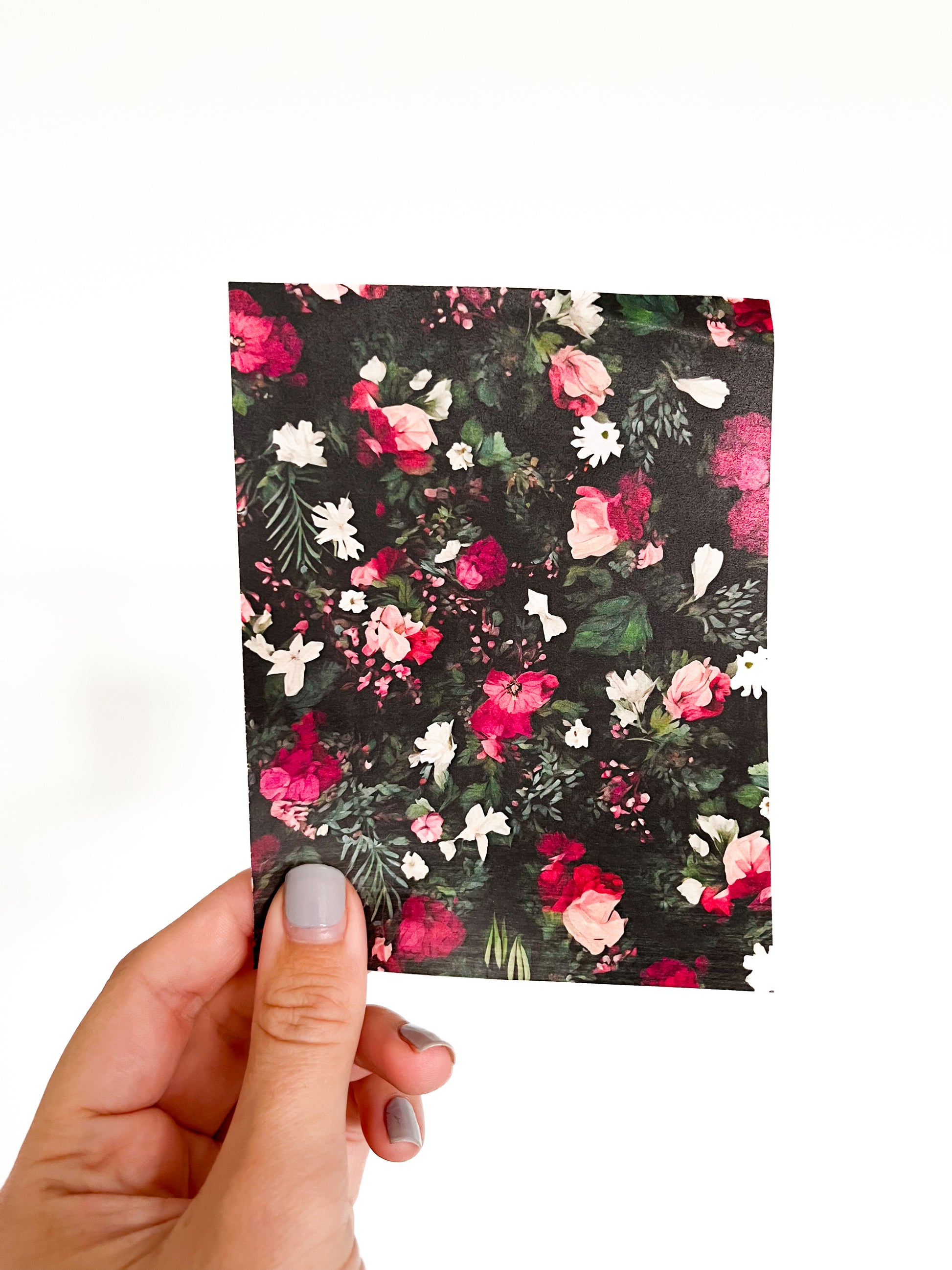 A hand with light gray painted nails is holding a rectangular piece of the Winter Garden Transfer by Agave Designs, featuring pink, red, and white flowers with green leaves on a black background. The backdrop is plain white.