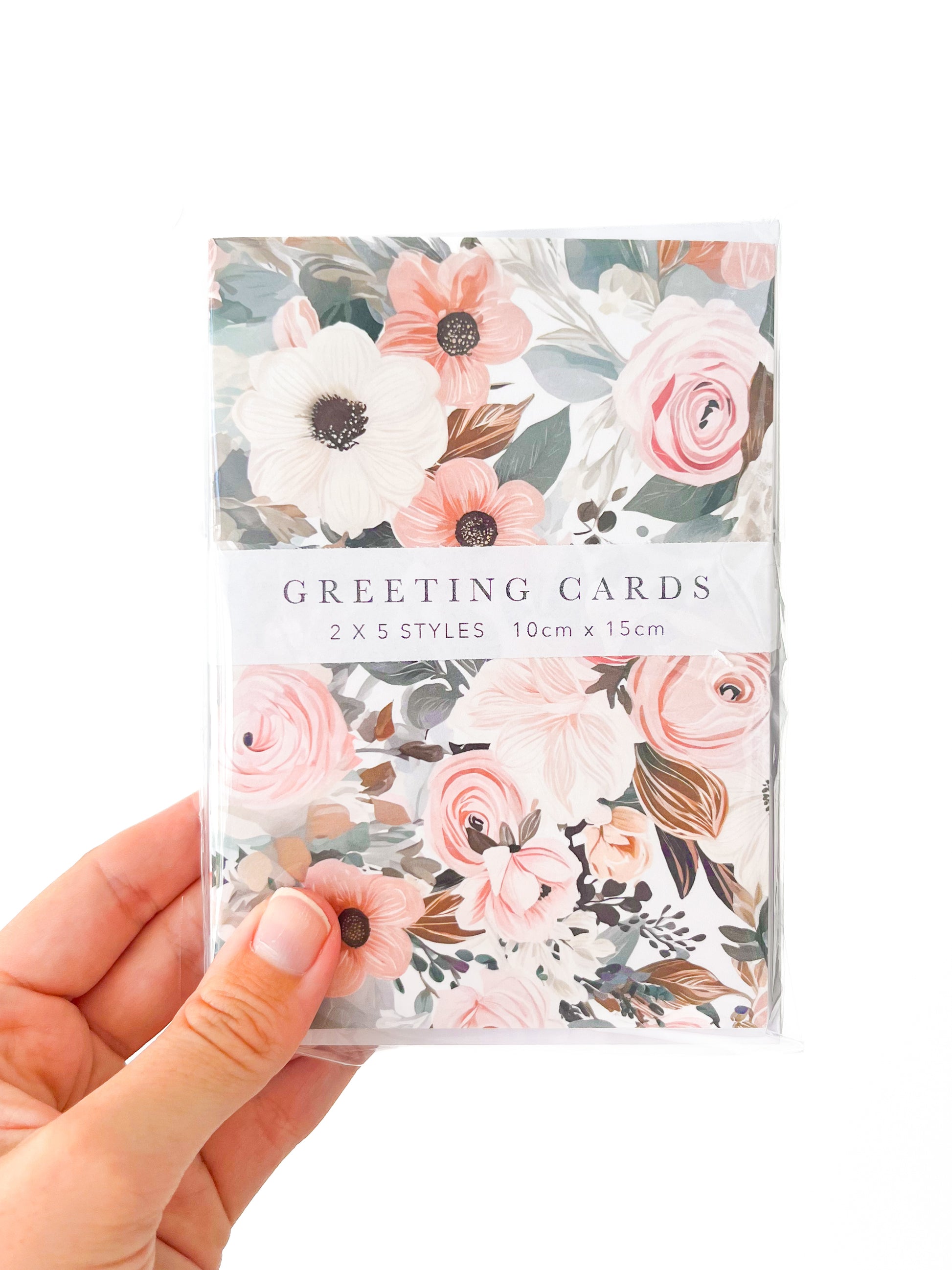 A hand holds a packet of Agave Designs greeting cards against a white background. The cards feature floral designs with pink and white flowers accented by green leaves. The packaging label reads: "GREETING CARDS, 2 x 5 STYLES, 10cm x 15cm." Next to the hand, there are delicate polymer clay earrings that beautifully complement the floral theme.