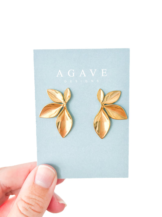 A hand holds a light blue card displaying golden leaf-shaped earrings from the brand "Agave Designs." These 1/2 Floral Studs are crafted from stainless steel and showcase a symmetrical design featuring two large leaves and a smaller leaf on each side, measuring 36mm x 24mm.