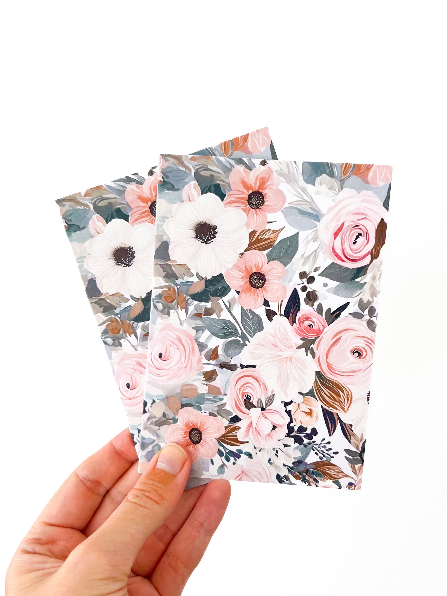 A hand holds three greeting cards with floral covers from Agave Designs against a white background. The covers feature an intricate design of various flowers, including white and pink blossoms with green leaves, reminiscent of the delicate charm found in polymer clay earrings. The overall aesthetic is delicate and elegant.