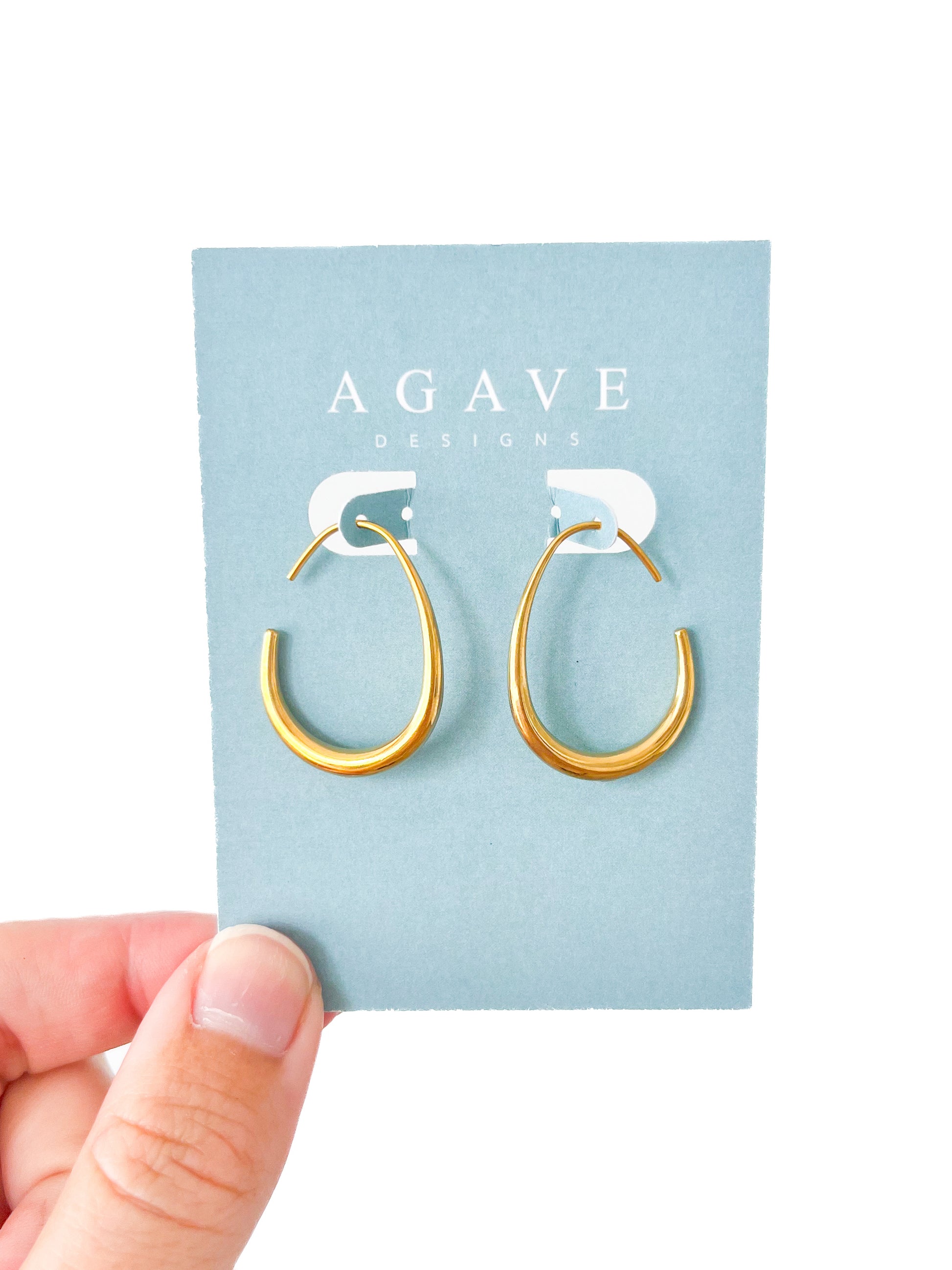 A hand holds a card with "AGAVE DESIGNS" printed on it. The card features "Oval Drops - Small," a pair of gold semi-open hoop earrings measuring 30mm x 22mm, against a light blue background. The earrings boast a sleek and minimalist design, crafted from stainless steel.
