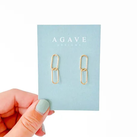 A hand with painted nails holds a blue card with two link earrings attached, plated in 14k gold. The card reads "Agave Designs" and features their product, the Double Chain. The background is plain white.
