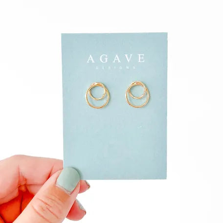 A hand with light green painted nails holds a blue card displaying a pair of Double Circle Gold Studs from Agave Designs. The background is plain white.