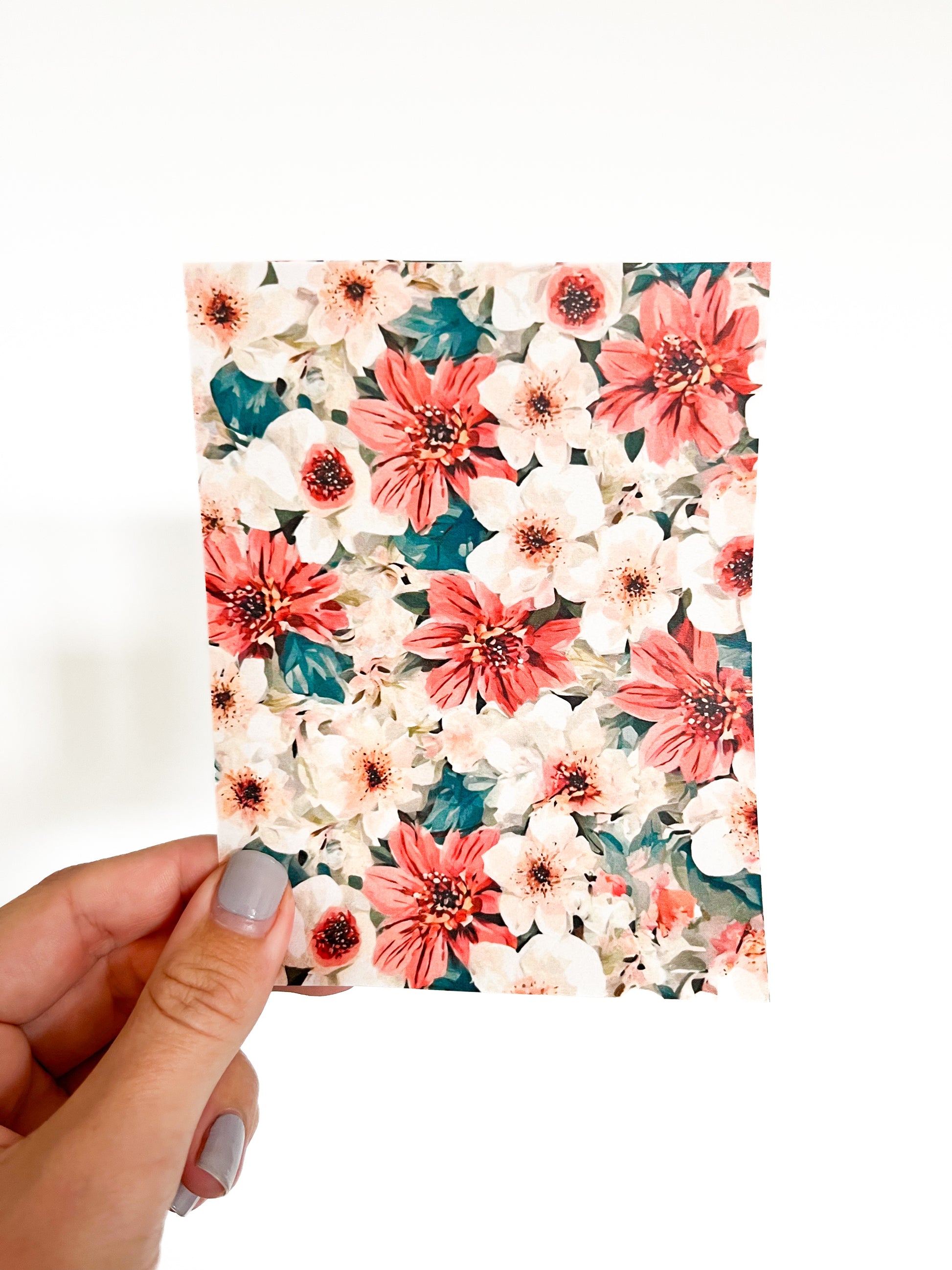 A hand with gray-painted nails holds a piece of July Transfer sheet by Agave Designs. The sheet showcases an assortment of red, pink, and white flowers with green leaves against a white background. The image is well-lit and has a minimalist style.