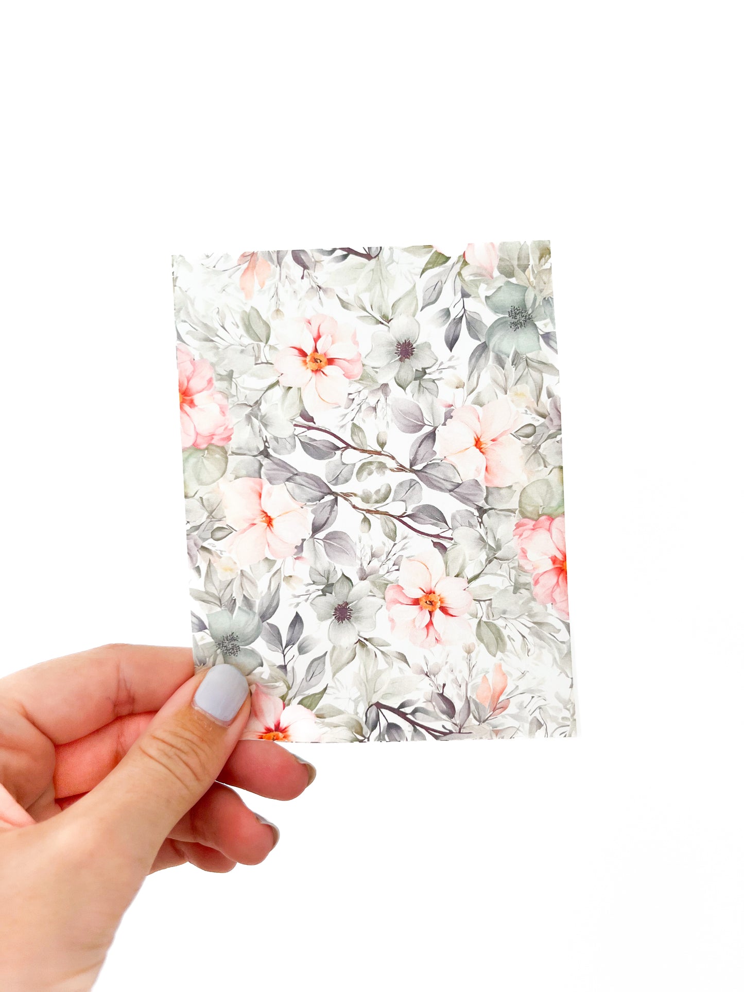 A hand with light-colored nails holds a small rectangular sheet of June Transfer sheet by Agave Designs, featuring a floral design with pink, white, and gray flowers accompanied by green leaves on a white background. The backdrop of the image is plain white.