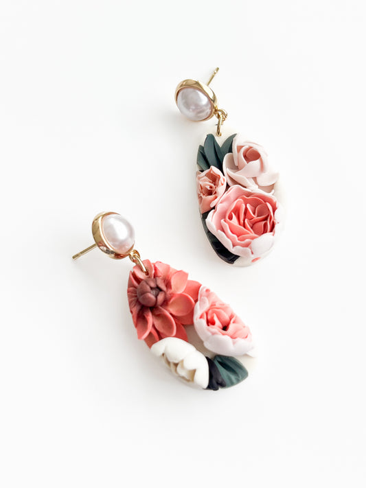 Introducing Iris by Agave Designs: A pair of elegant drop earrings featuring gold-plated studs with white pearls. The hanging portion, measuring 60 x 30mm, is adorned with intricate, colorful floral designs in shades of pink, peach, and white, surrounded by green leaves on a forest green background.