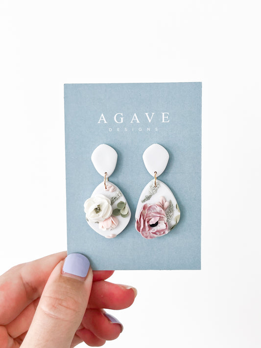 A hand with lavender nail polish holds a light blue "Agave Designs" card displaying the Julie earrings—white teardrops featuring delicate 3D floral designs in soft pink and cream.