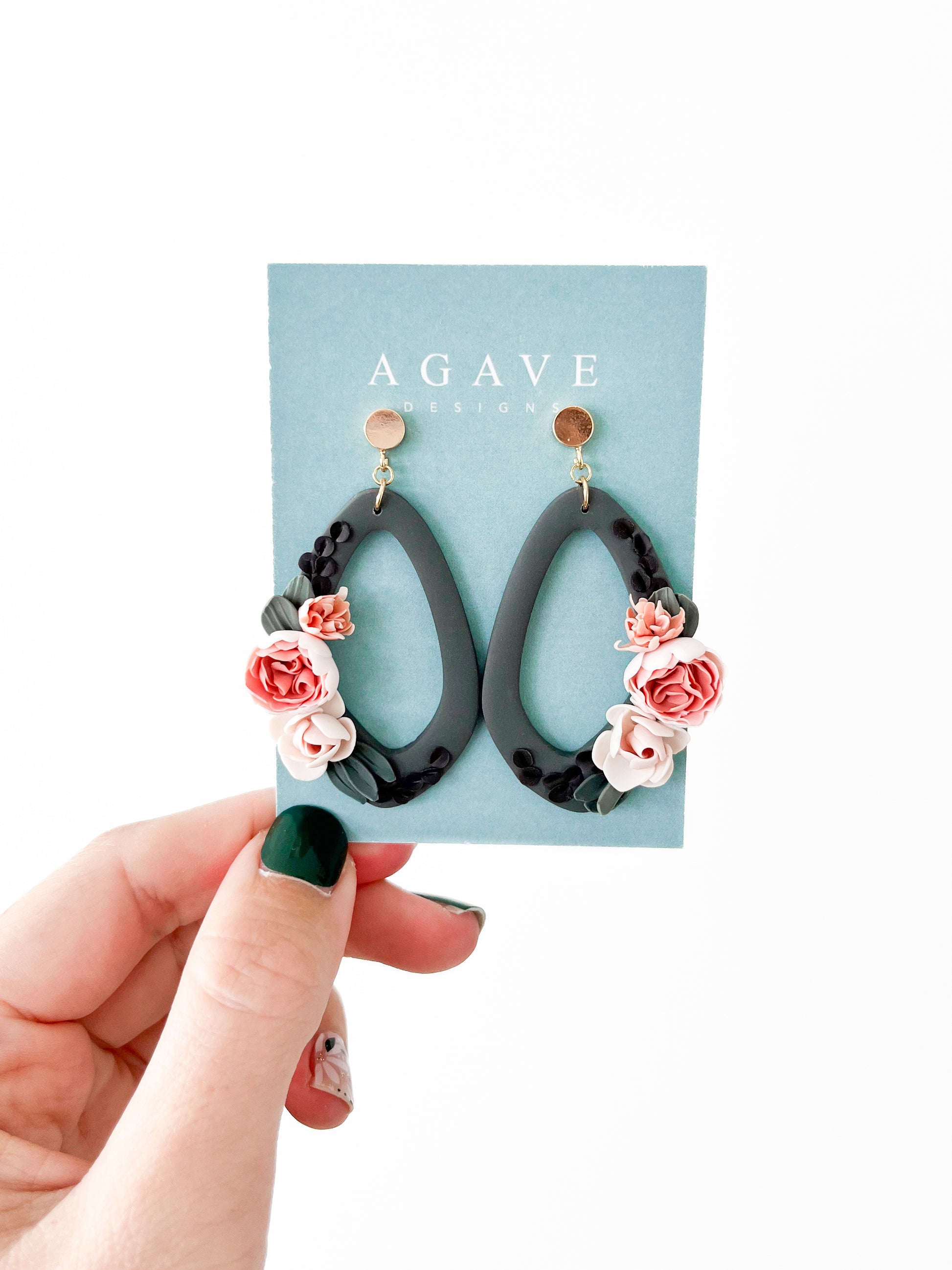 A hand with forest green nail polish holds a pair of black teardrop-shaped earrings, named Harmony, adorned with small, intricate pink and white flowers. The 60 x 30mm earrings are mounted on a light blue card displaying the text "Agave Designs.