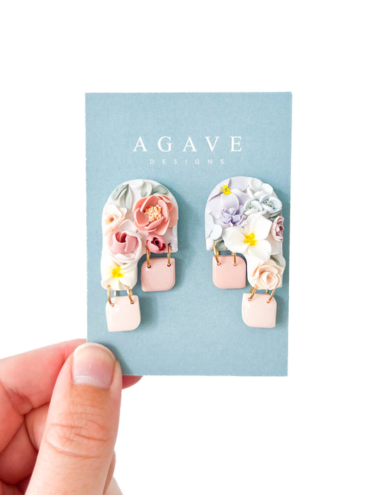 A hand holds a card with "Agave Designs" on it. Attached to the card is a pair of Lisa made-to-order sterling silver earrings with an arch shape, adorned with intricate pastel-colored flowers, and pink rectangular dangles at the bottom. The background is white and minimalistic.