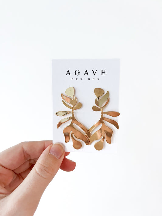 A hand displays a card with the "Agave Designs" brand, presenting exquisite gold-toned geometric leaf danglies made from high-quality zinc alloy against a pristine white background.