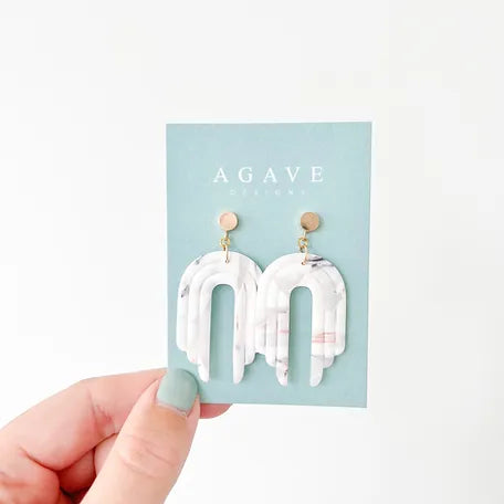 A hand with mint green nail polish holds a card displaying a pair of 55mm x 28mm white and marbled dangle earrings. The card has a light blue background with the brand name "Agave Designs" written on it. The gold-plated earrings feature a modern, arched design and are named Fay.