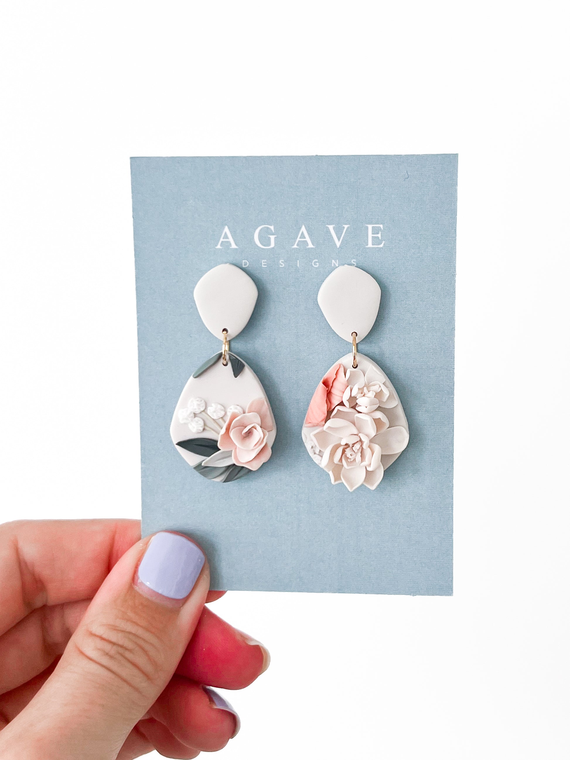 A hand with lavender nail polish holds a blue "Agave Designs" card showing the Tammy earrings: 40mm x 22mm sterling silver teardrop earrings with raised floral designs in soft pink, white, gray, and sage.