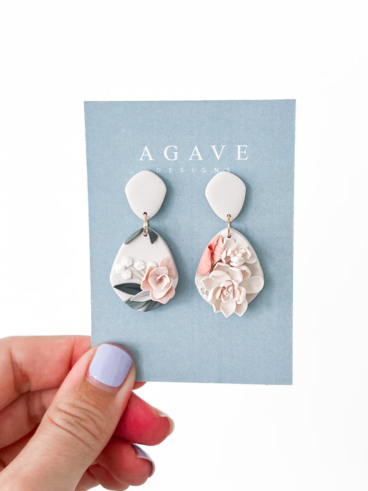 A hand with lavender nail polish holds a blue "Agave Designs" card showing the Tammy earrings: 40mm x 22mm sterling silver teardrop earrings with raised floral designs in soft pink, white, gray, and sage.