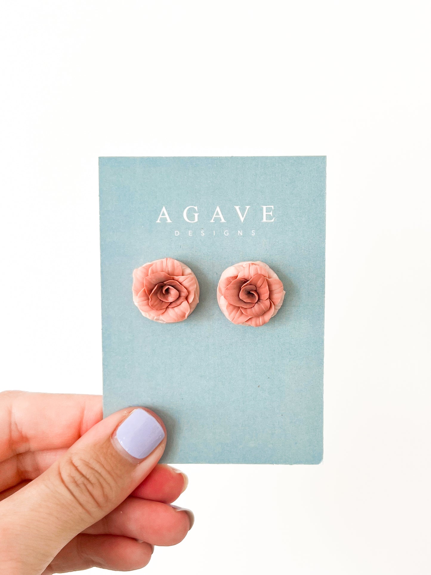 A hand with light purple nail polish holds a blue Agave Designs card displaying 20mm coral pink Rose studs crafted in sterling silver.