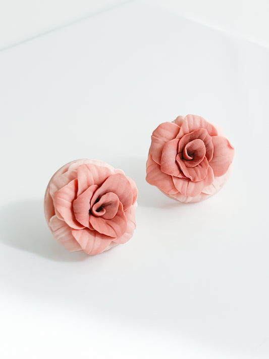 Two coral pink rose-shaped Rose studs by Agave Designs, displayed on a clean white surface with soft lighting.