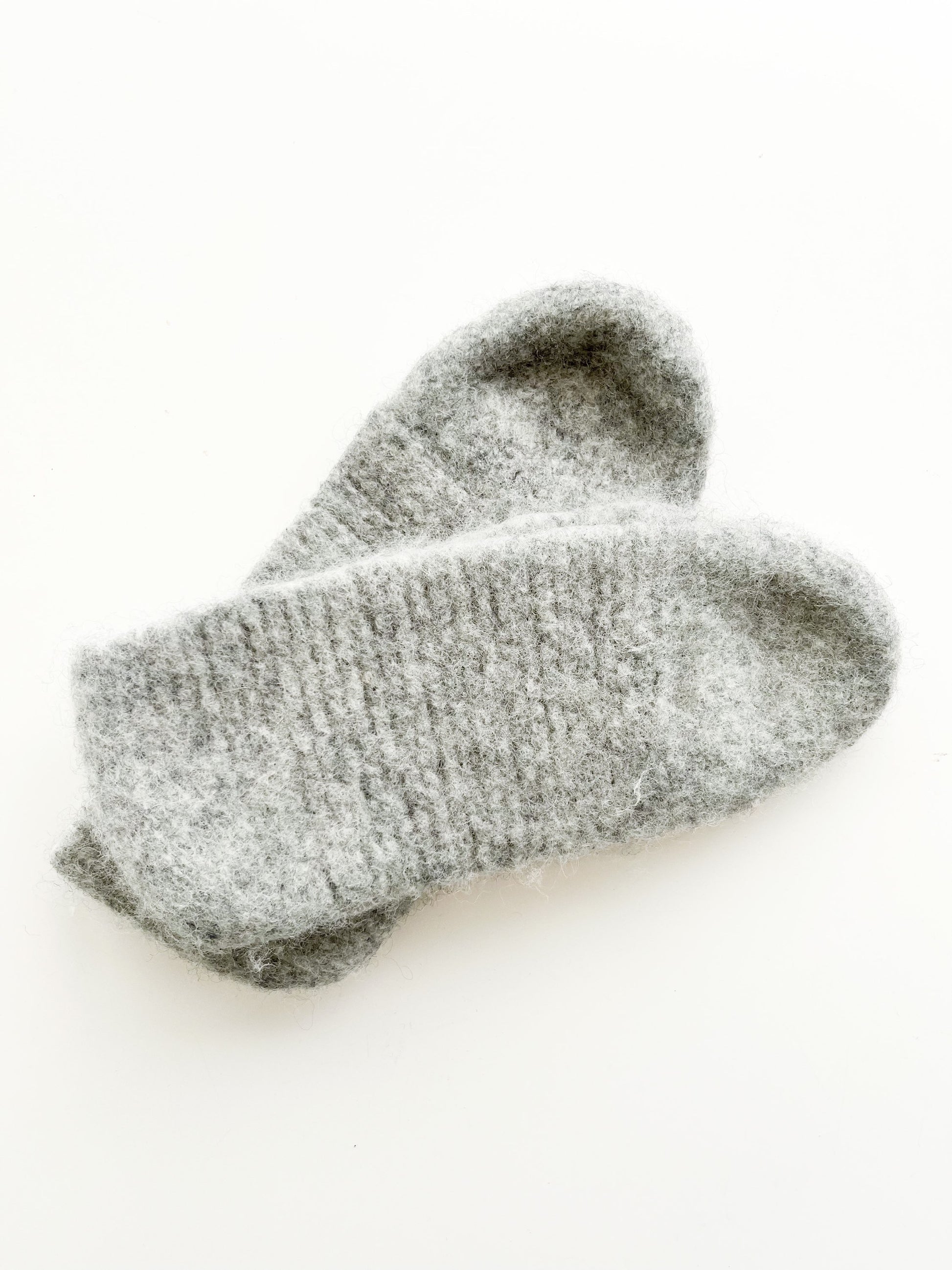 A pair of light grey wool slippers from Agave Designs, made with Norwegian wool from Sandes Garn, lying flat on a white surface. The slippers appear thick and fuzzy, suggesting warmth and comfort. For best results, hand wash them to maintain their cozy texture.