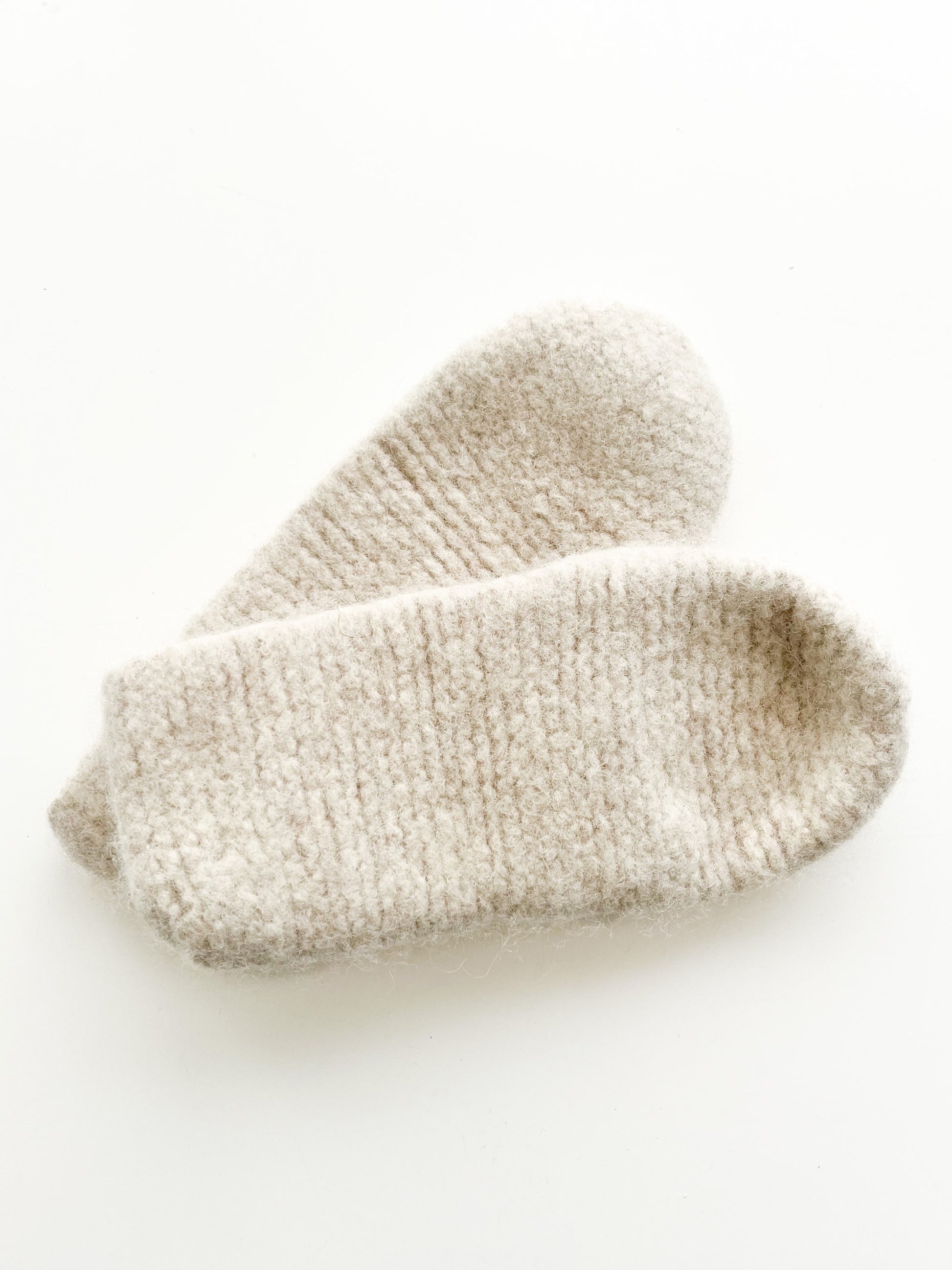 An Oatmeal Wool Slipper, crafted by Agave Designs using high-quality Norwegian wool, laying crossed over each other on a white background. The slippers have a soft and cozy texture that suggests warmth and are made to order.