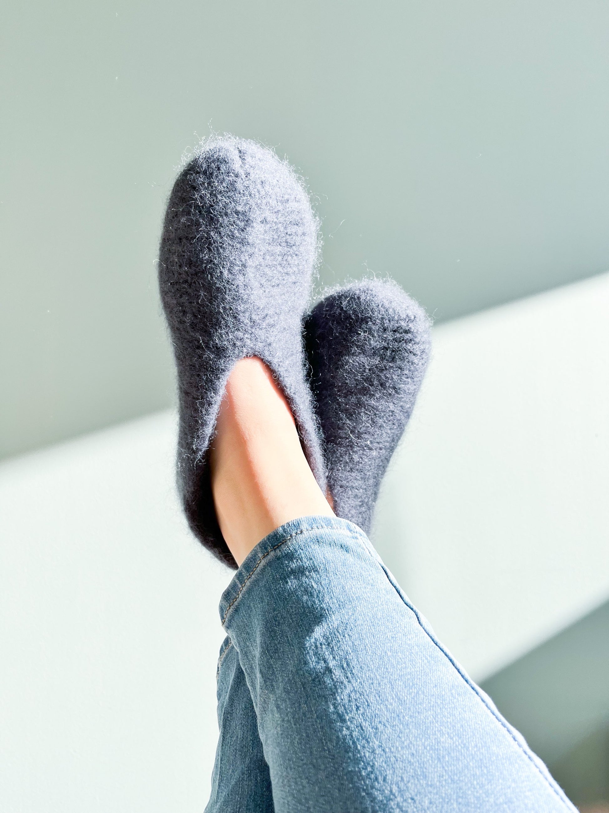 A person wearing blue jeans is relaxing with their legs crossed, showcasing cozy, fuzzy Soft Navy Wool Slippers by Agave Designs against a light background. Their feet are slightly elevated and the natural light highlights the soft texture of the slippers.
