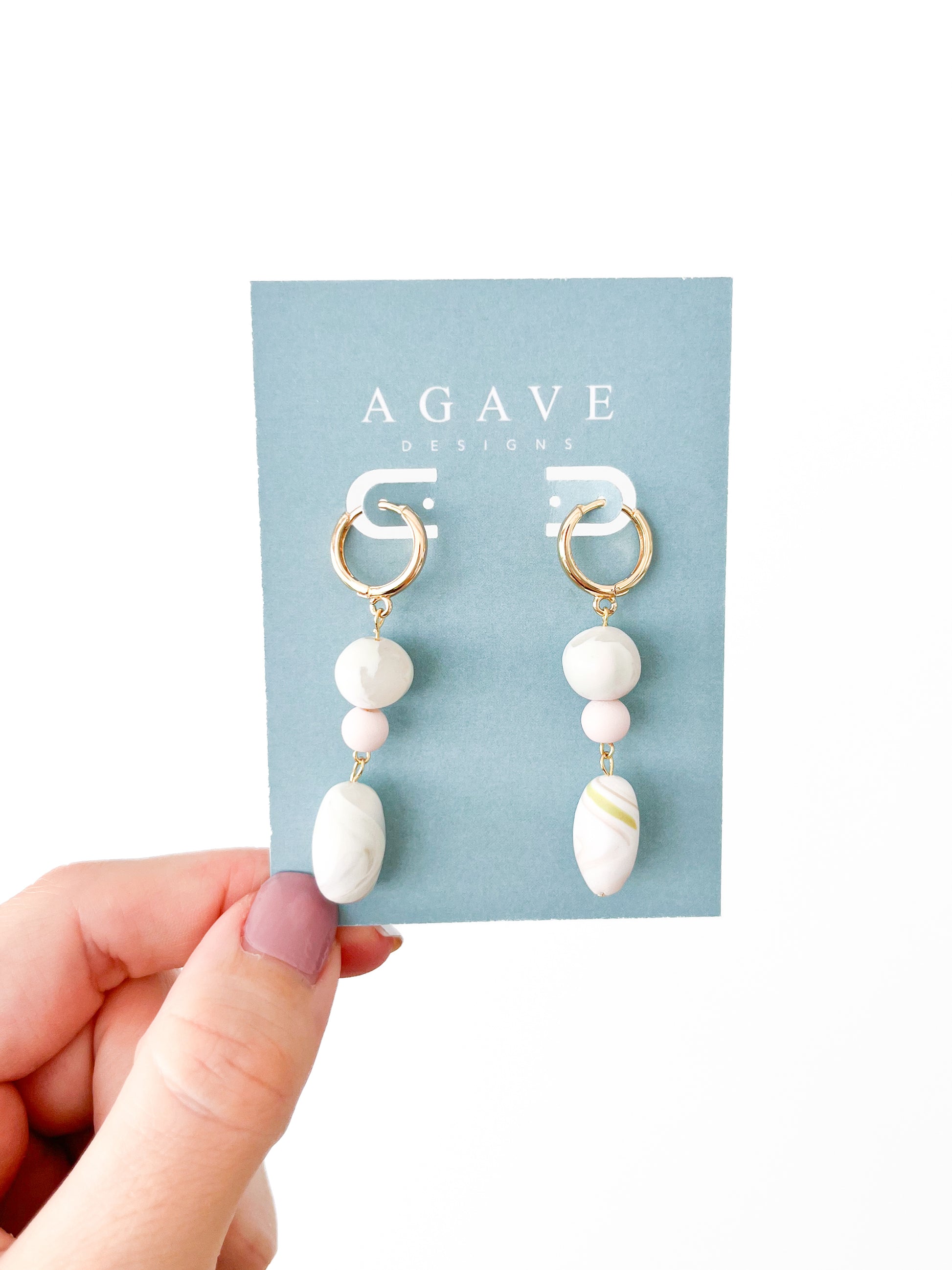 A hand with light purple painted nails is holding a blue card labeled "Agave Designs" showcasing a pair of made-to-order Olivia earrings. The earrings feature gold plated hoops, white bead dangles, pink accents, and teardrop-shaped white marbled pendants measuring 53mm x 10mm.