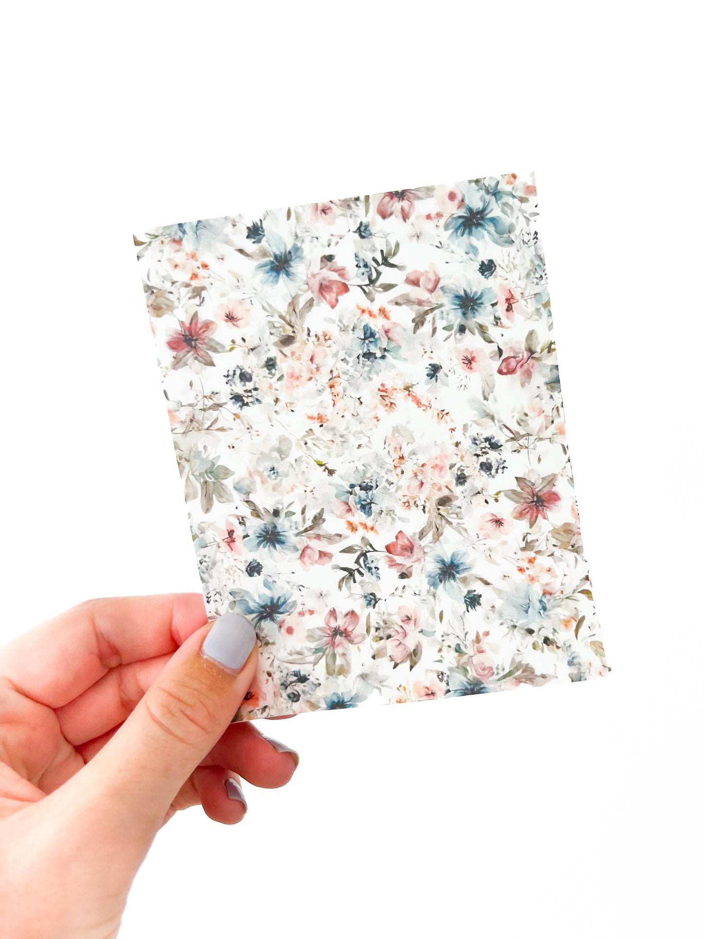 A hand with light-colored nail polish holds the Agave Designs "March Transfer Sheet," a small rectangular piece of transfer paper adorned with a floral design featuring various pastel-colored flowers on a white background. The image is set against a plain white backdrop.