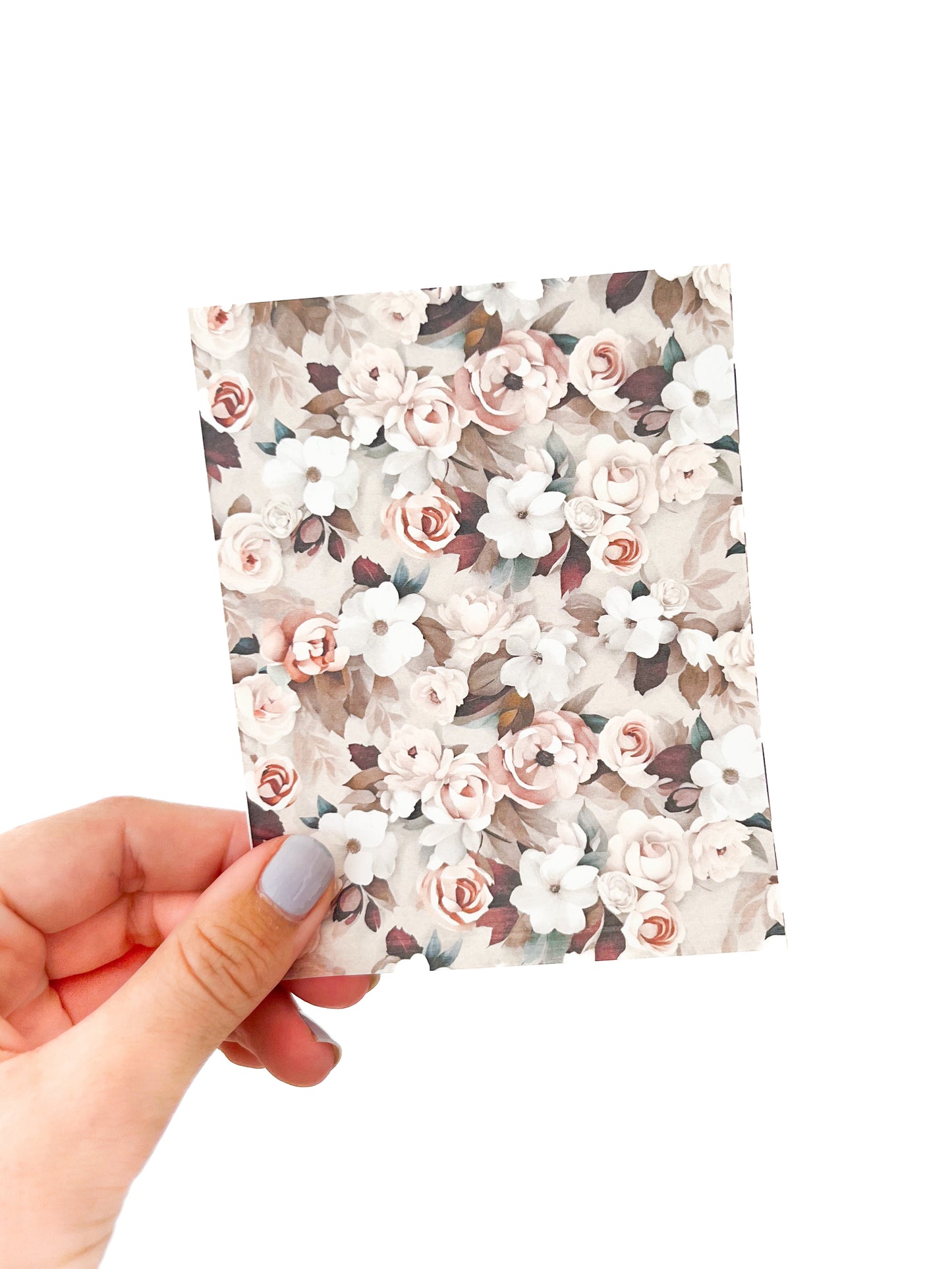 A hand with light-colored nail polish holds a small rectangular sheet of February Transfer Sheet by Agave Designs. The sheet is adorned with various flowers in shades of pink, white, and beige, set against a light background.