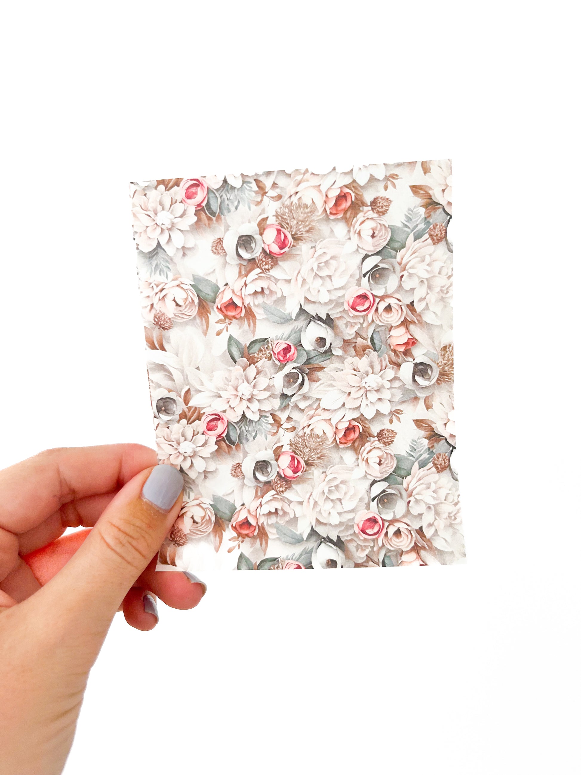 A hand with light blue painted nails holds a rectangular January Transfer Sheet from Agave Designs. The sheet features a floral pattern showcasing an array of flowers in shades of white, pink, and beige on a white background. The photo is taken against a plain white backdrop.