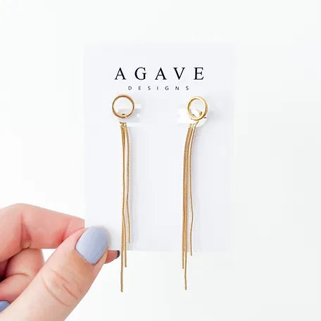 A hand with light purple nail polish holds a white card displaying a pair of Gold Danglies from Agave Designs. The earrings, measuring 85mm x 10mm, feature a small circular stud and multiple dangling chains. The card has the text "Agave Designs" printed at the top.